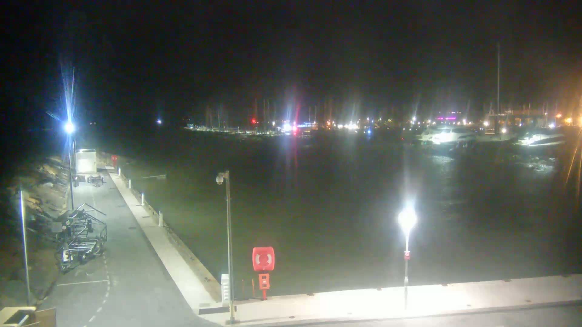 A nighttime view of a marina with boats docked, illuminated by several lights, with somewhat choppy water.