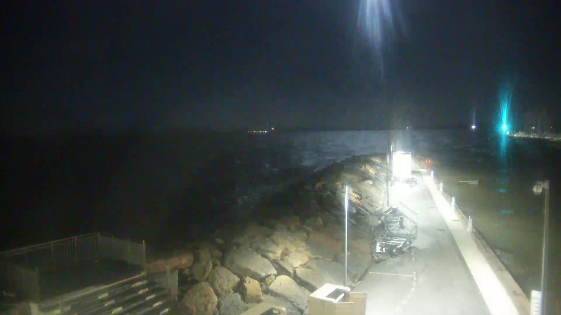 A nighttime coastal scene shows a rocky breakwater next to a paved walkway, with dark, choppy water visible beyond.