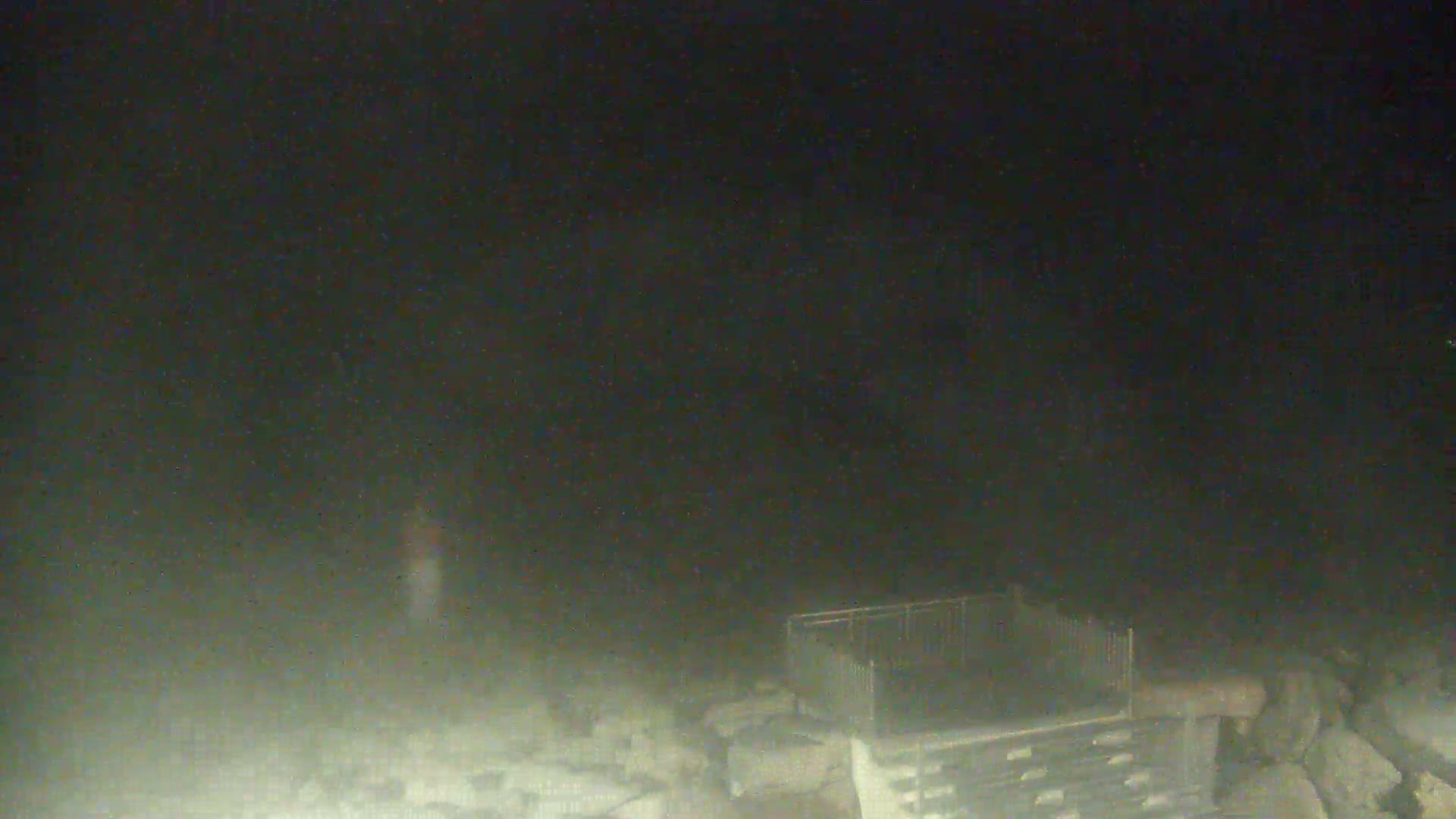 A dark, hazy night scene shows a metal structure resembling a gate or fence near a pile of rocks.