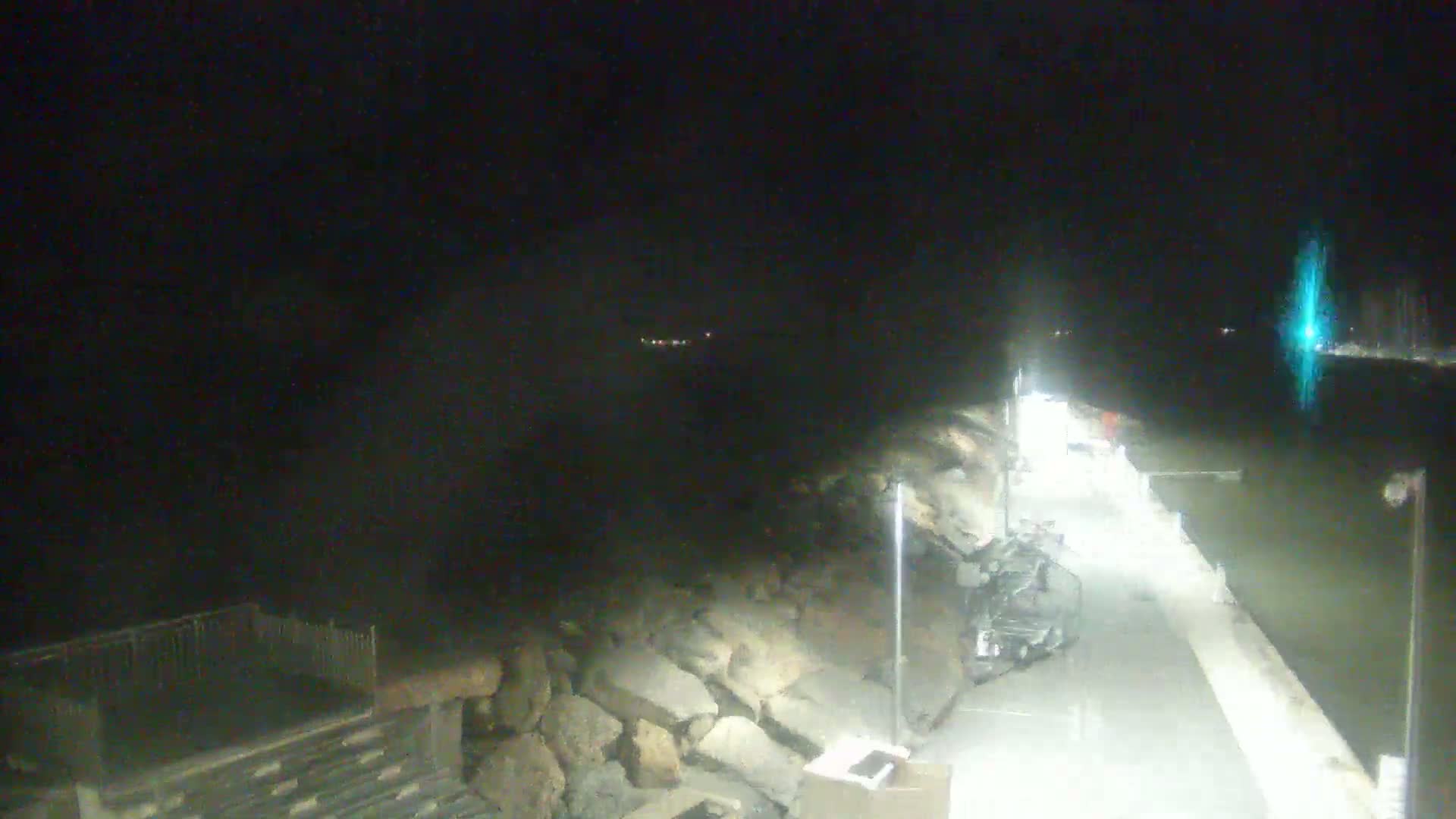 A nighttime view of a paved walkway along a rocky shoreline with a body of water visible in the background.