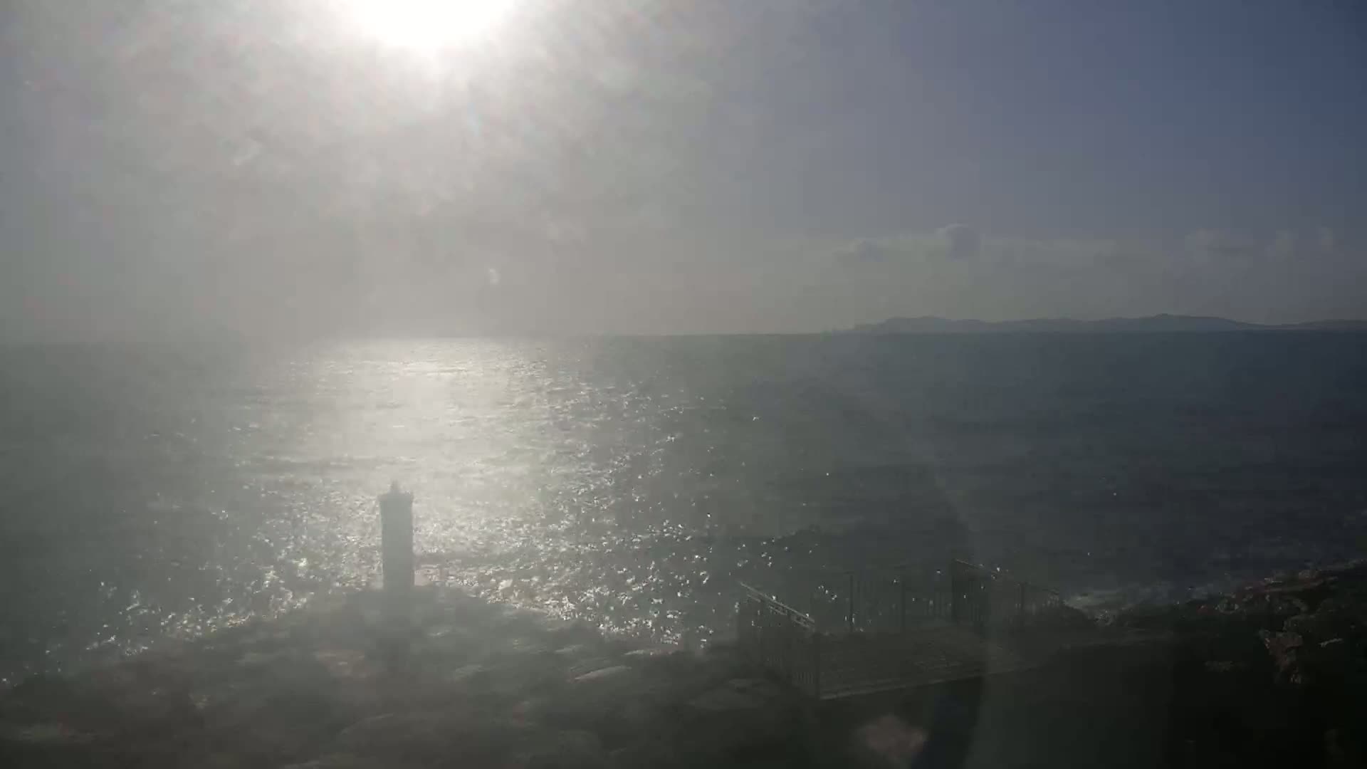 A hazy sun shines over a rocky coast and choppy sea, with a lighthouse and a railing structure visible in the foreground.