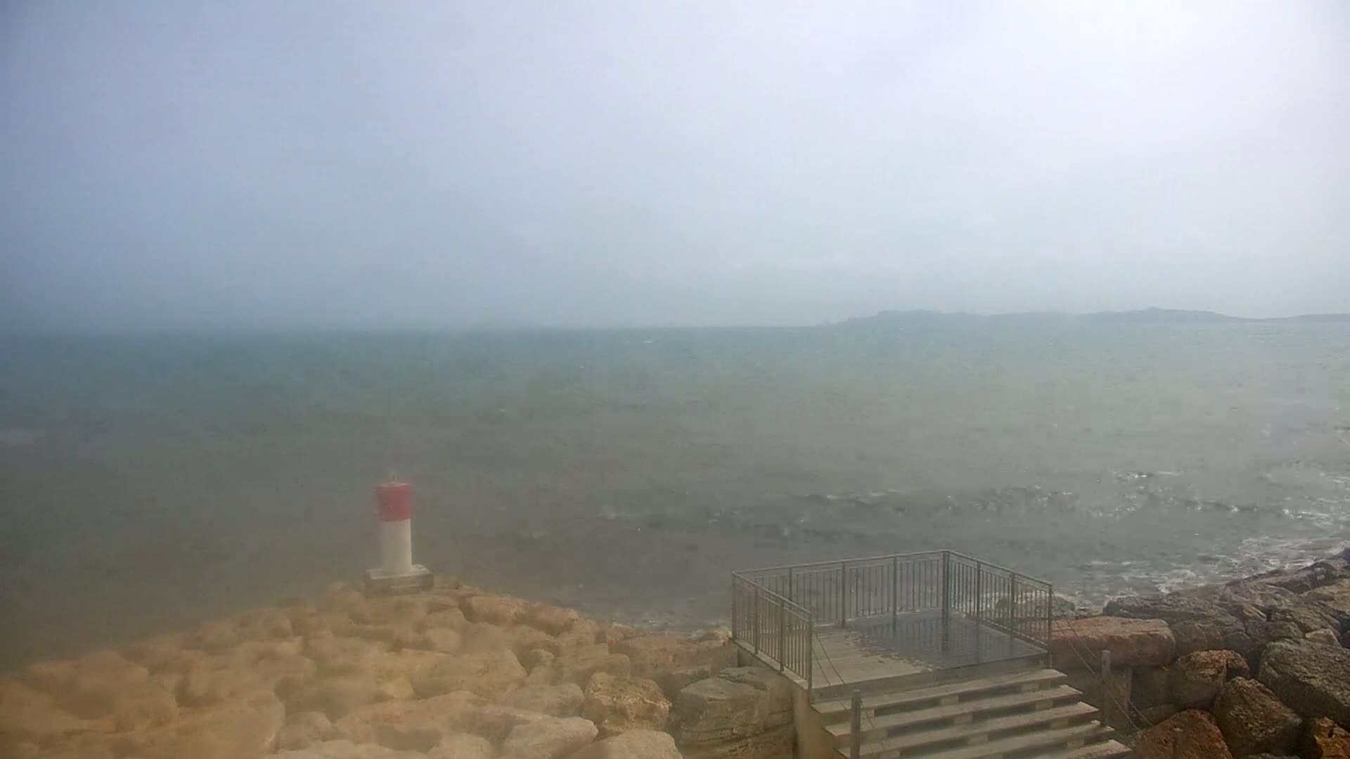 A rocky shoreline with a small lighthouse and a set of stairs overlooks a hazy, choppy sea on a foggy day.