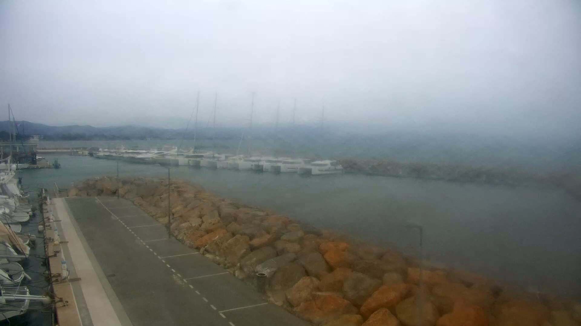 A foggy view of a marina with numerous boats moored, partially obscured by a rocky seawall and a paved area.