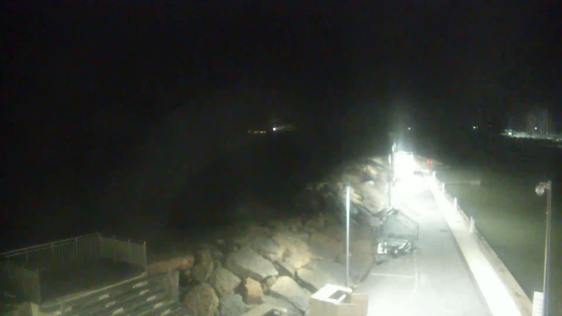 A nighttime, brightly lit pier extends over dark water, bordered by a rocky shoreline.