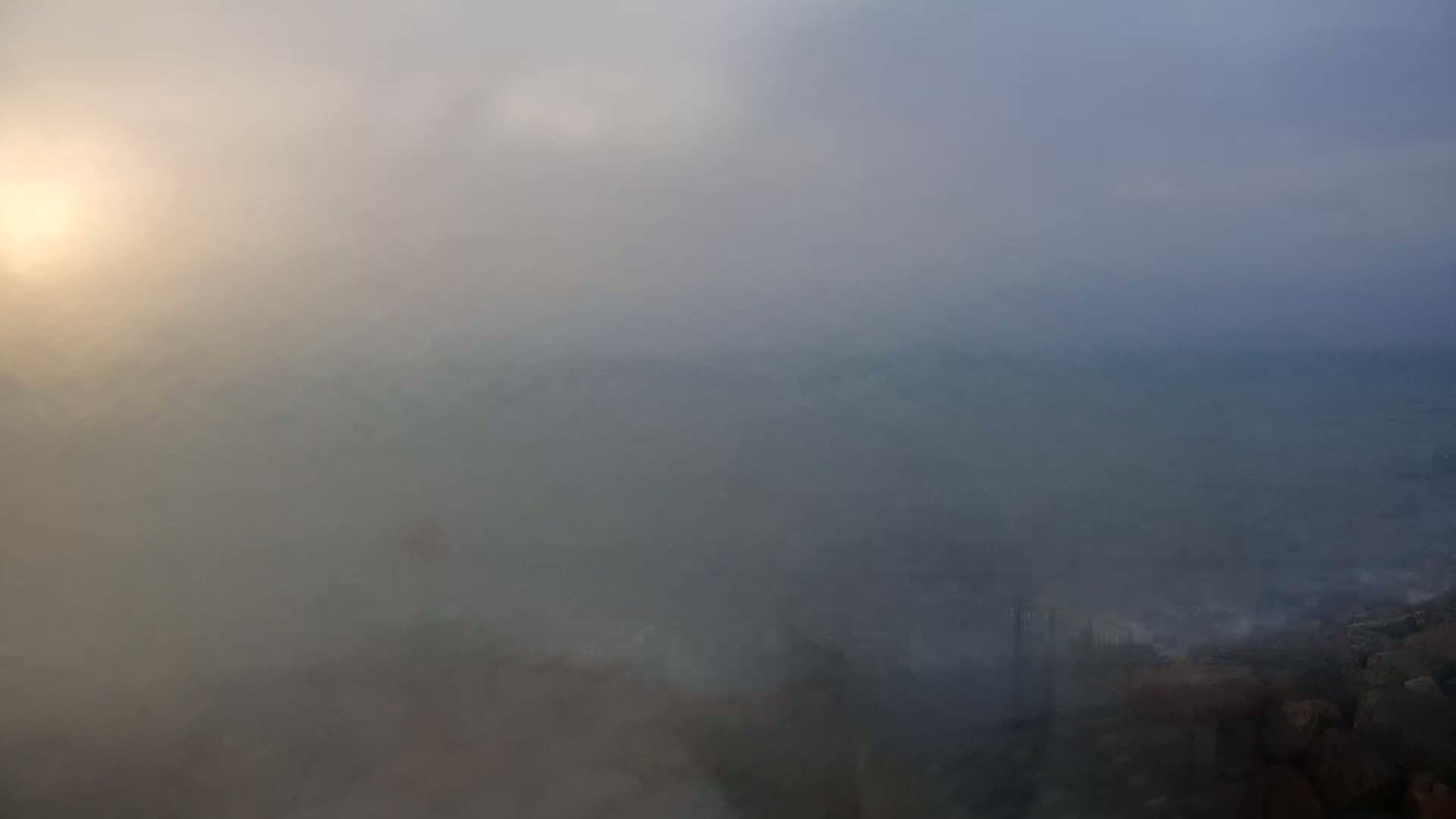 A hazy, sunlit landscape stretches out, shrouded in dense fog that obscures the details of the land and sea below.