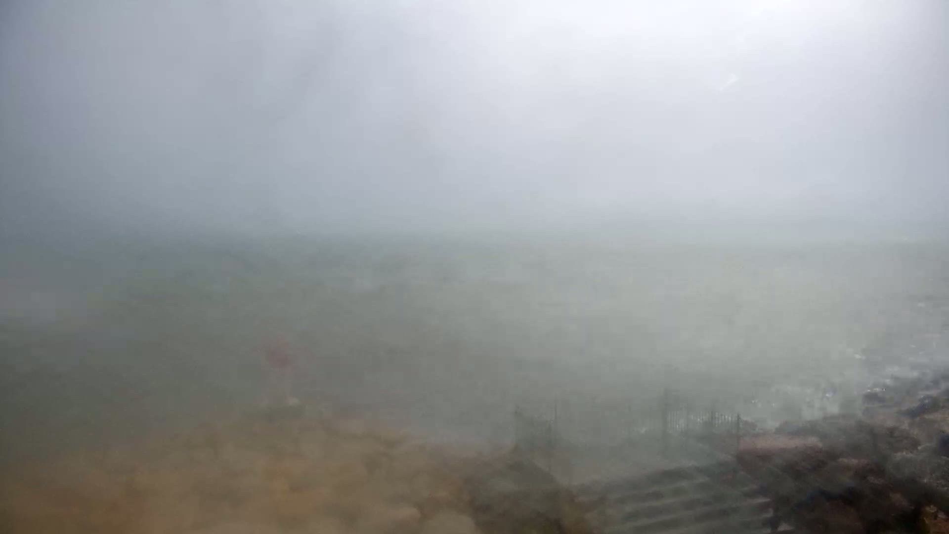 A misty, overcast scene shows a tan ground sloping down to a blurry, foggy expanse of what looks like water or a flat, wet land.
