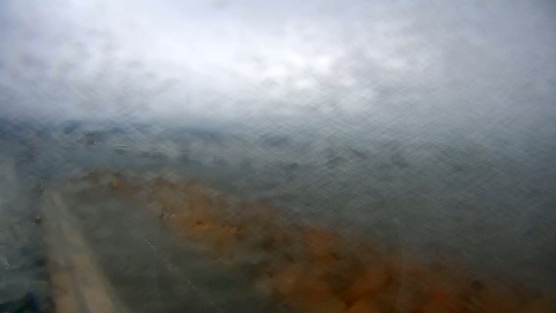 A blurry view of a land and water landscape seen through a rain-streaked window or similar surface, under an overcast sky.