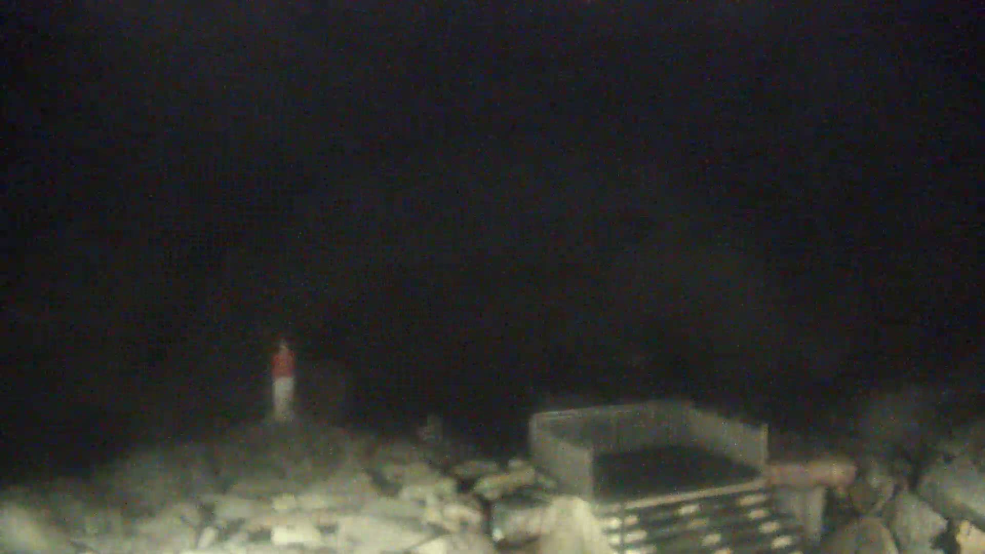 A blurry, nighttime image shows a person standing on a rocky, debris-strewn area near a dark, metal container.
