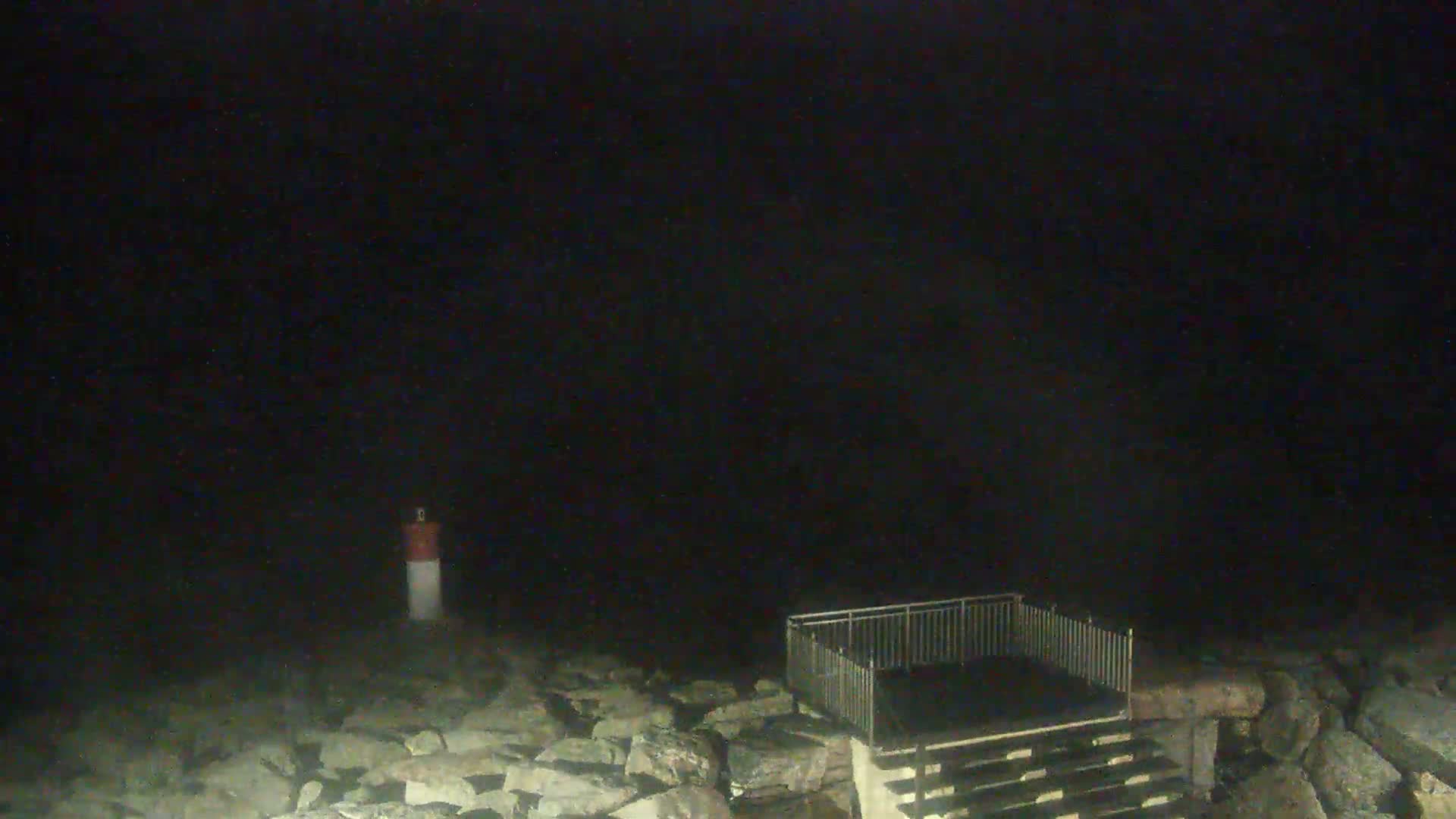 A small, fenced-in platform with stairs sits on a rocky shoreline at night, next to a small, red-and-white marker.