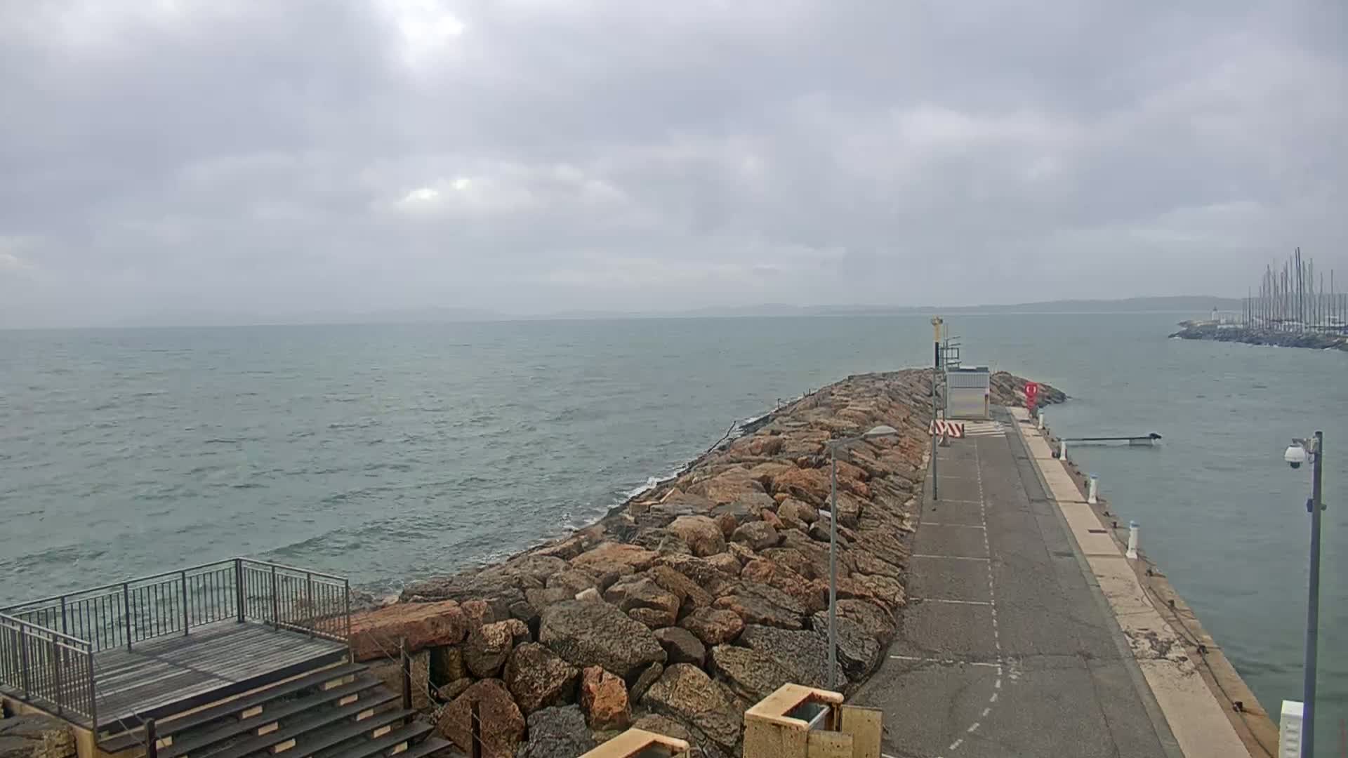A rocky breakwater extends from a paved walkway into a choppy sea under a cloudy sky.
