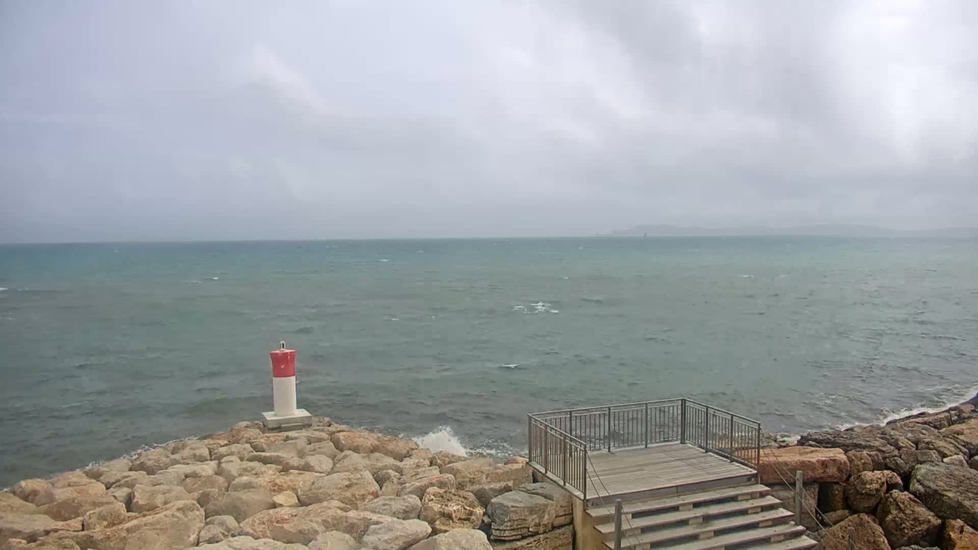 A rocky shoreline with a small viewing platform and a red-and-white marker is shown under a cloudy sky, with a choppy sea extending to the horizon.