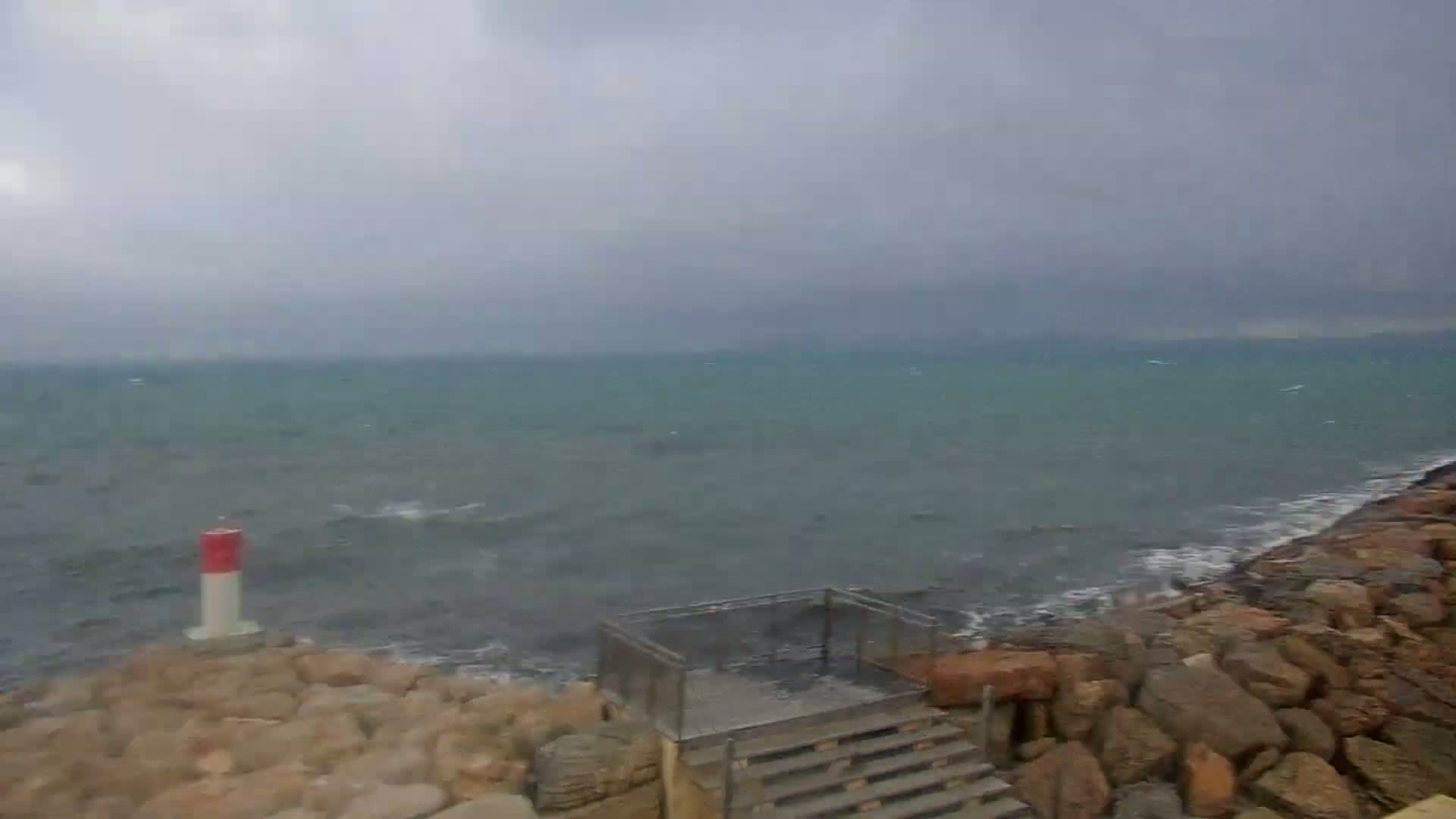 A rocky shoreline with a small staircase and a red and white marker is shown, with a choppy sea under an overcast sky.