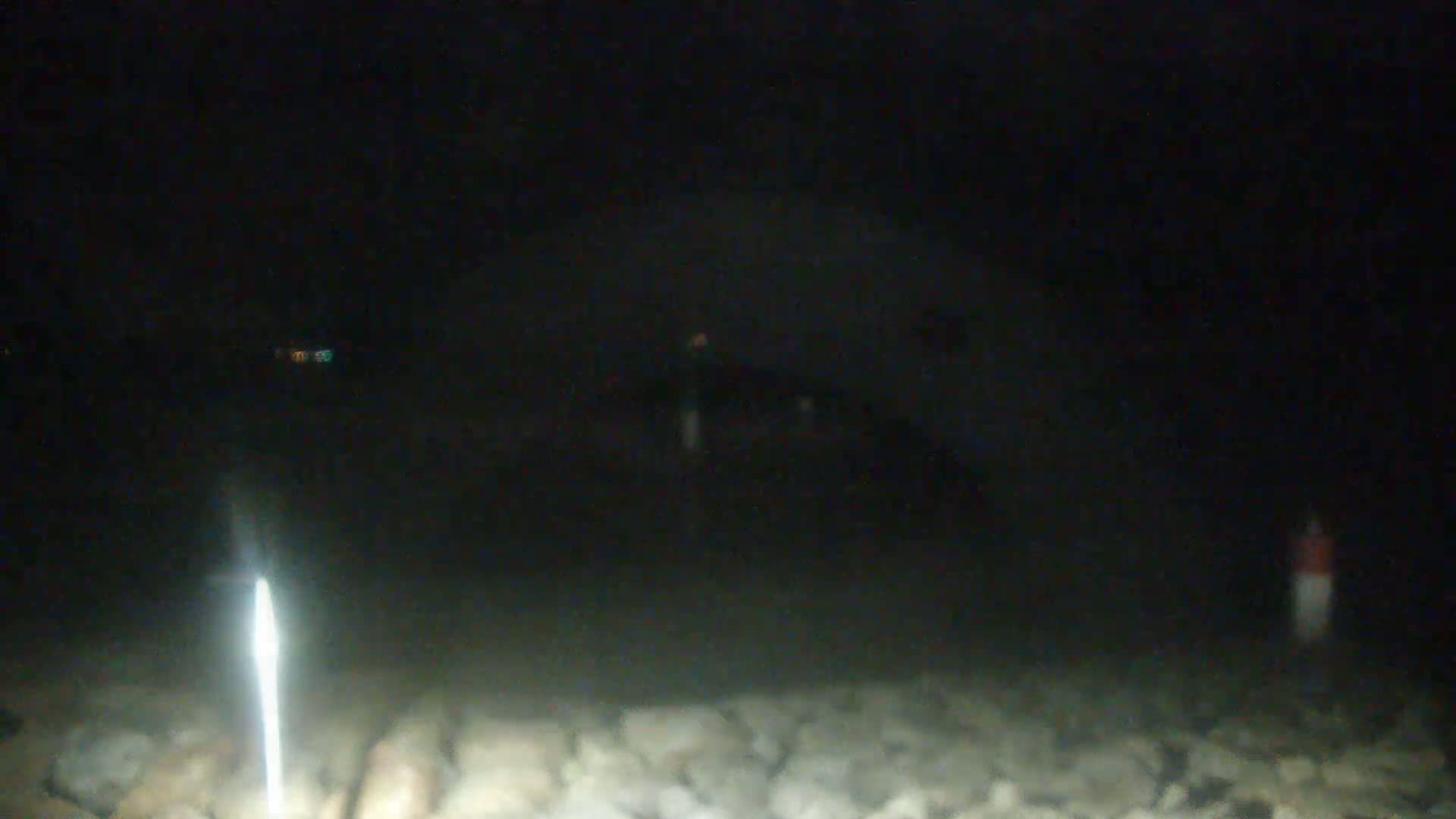 A blurry, nighttime image shows a dark structure in the distance, illuminated faintly, with a bright vertical light source in the foreground and a rocky area at the bottom.