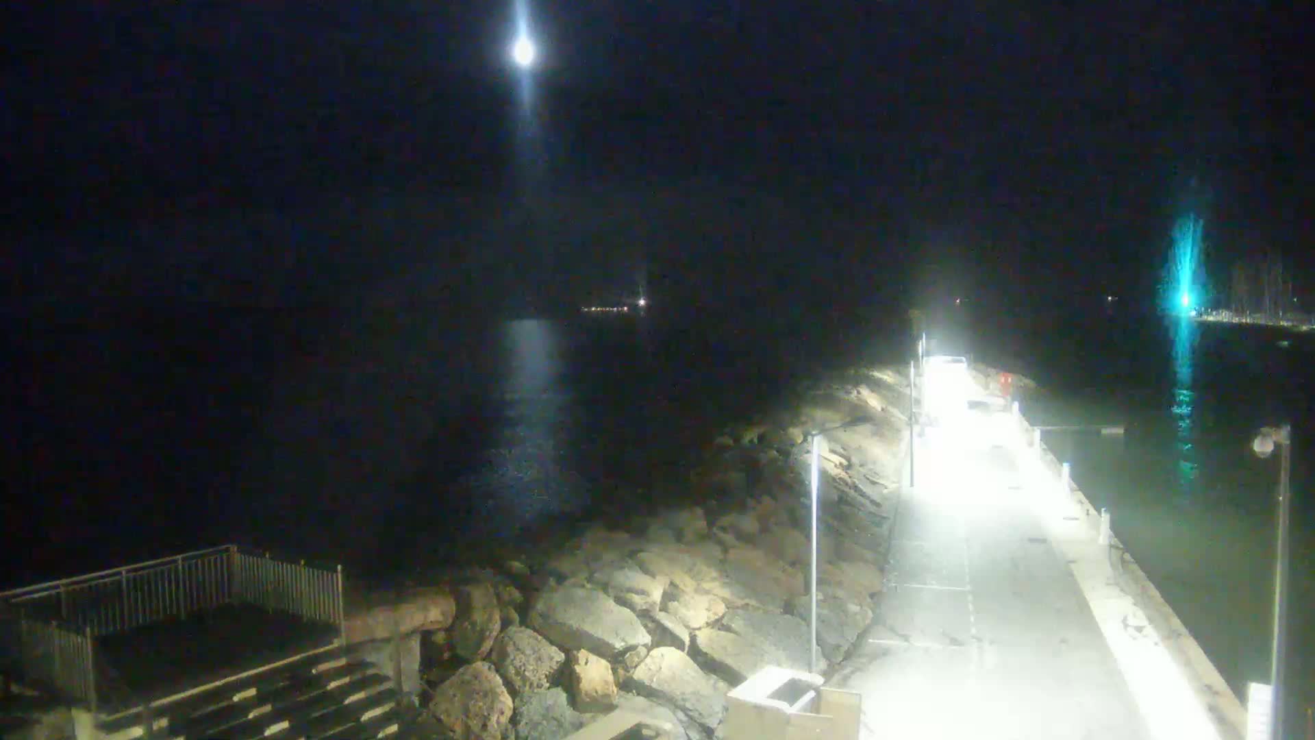 A nighttime view of a lit walkway along a rocky shoreline with a body of water showing a bright light source in the distance under a moonlit sky.