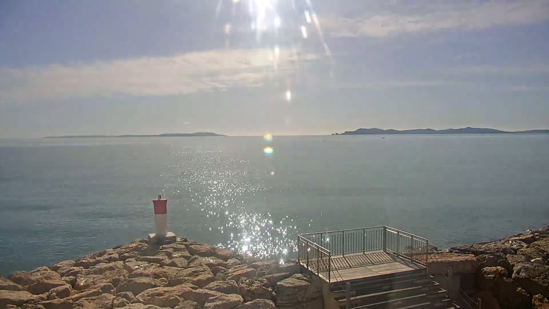 A rocky shoreline with a small railing and steps overlooks a calm, sunlit sea with distant islands under a partly cloudy sky.