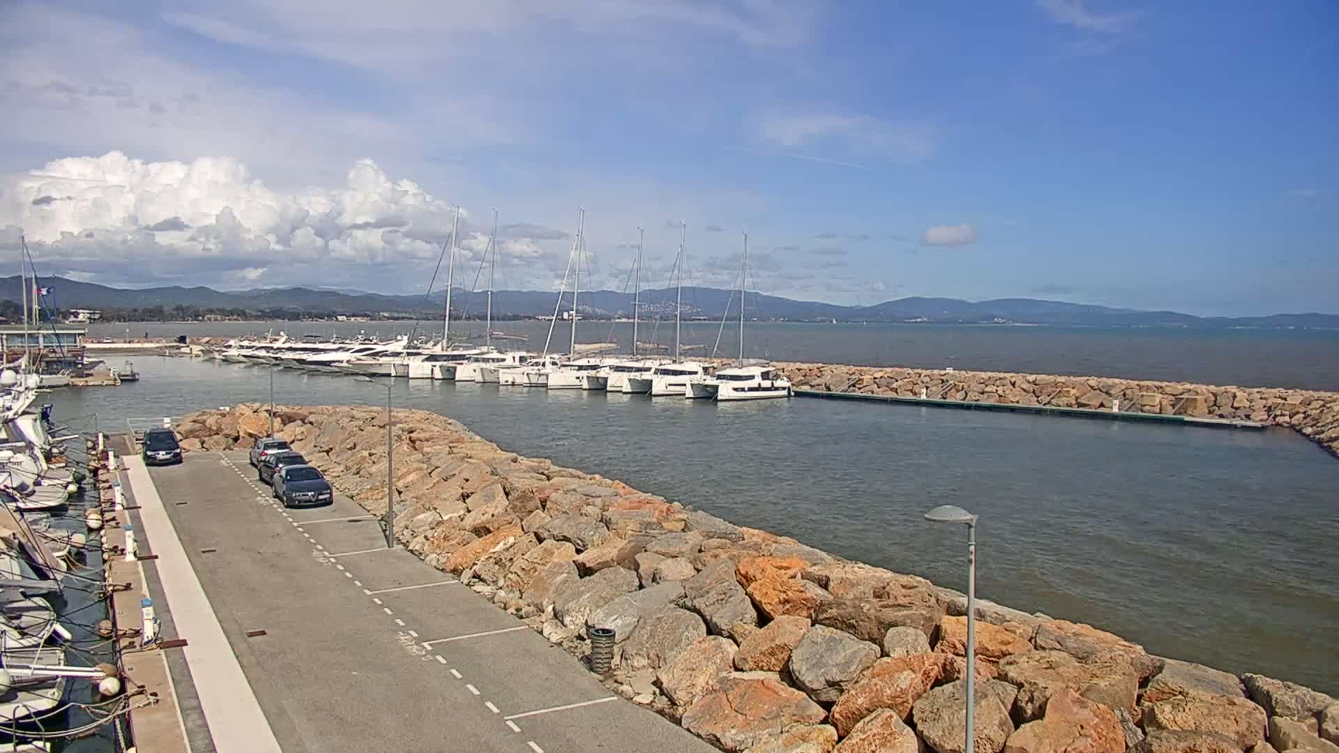A marina with numerous sailboats and motorboats docked, situated beside a rocky breakwater on a partly cloudy day.