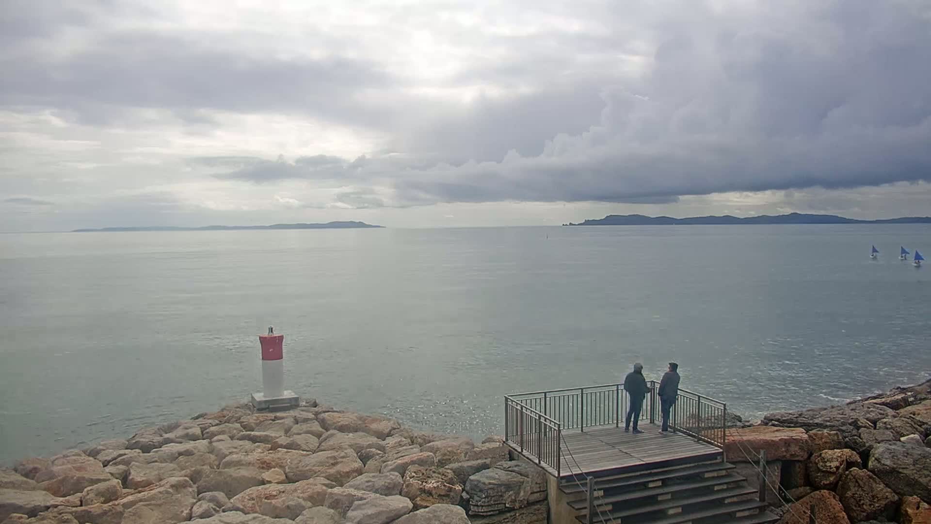 Two people stand on a wooden platform overlooking a calm sea under a cloudy sky, with a small lighthouse and distant islands visible.