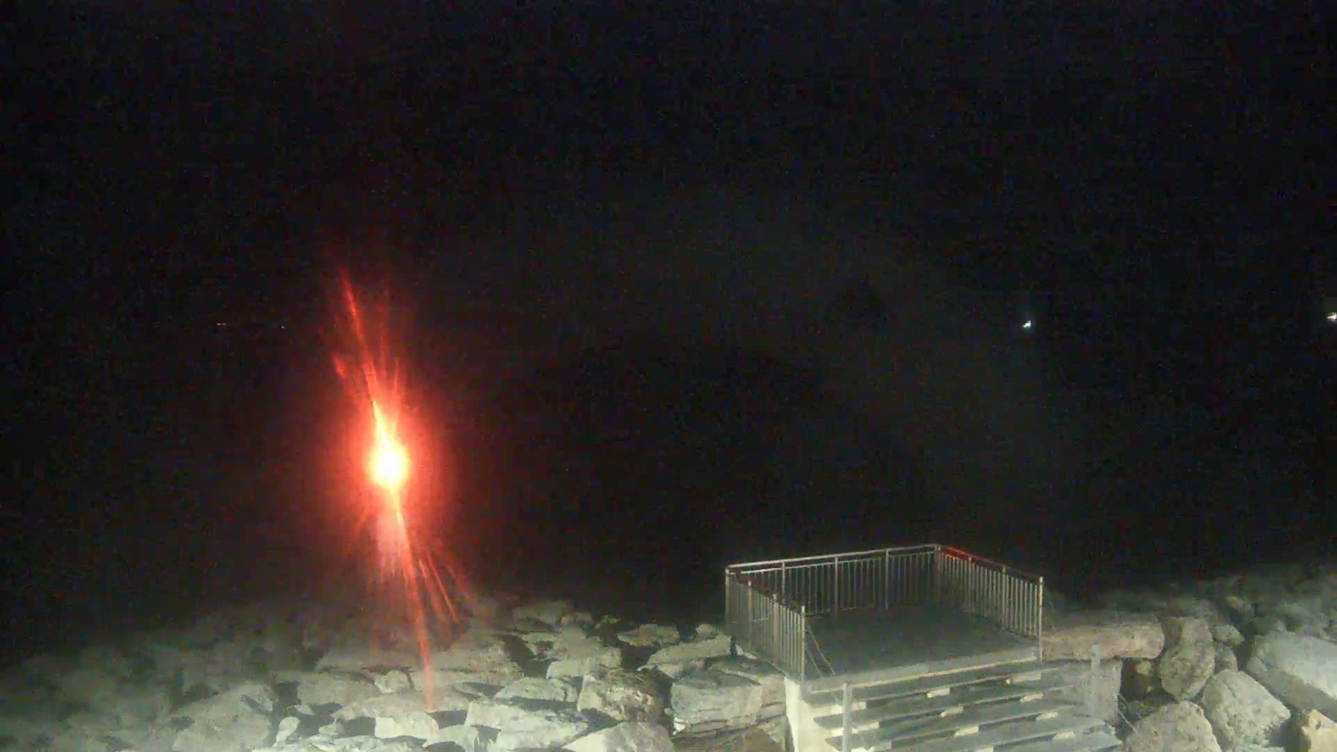 A bright red light shines over a rocky shoreline at night, next to a set of stairs leading to a metal platform.