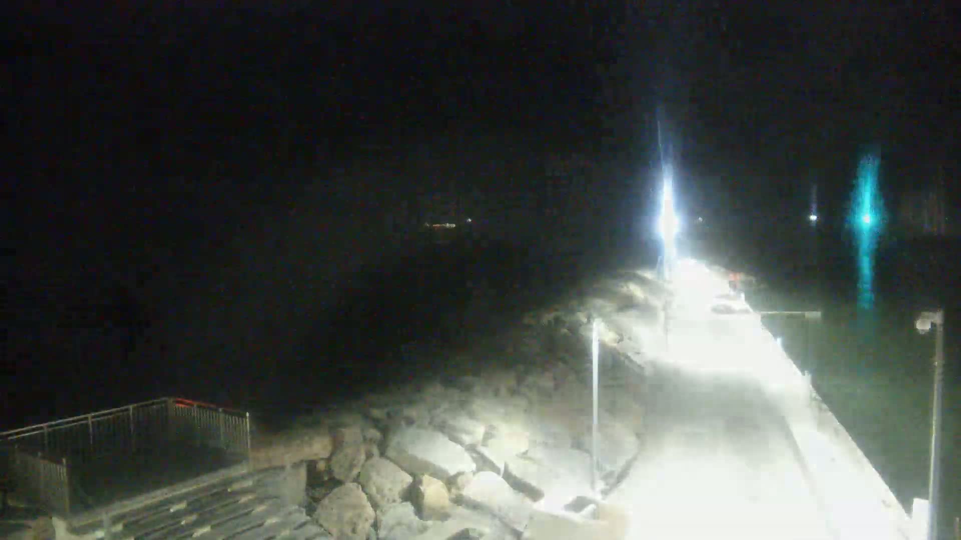 A nighttime view shows a brightly lit pier extending over dark water, with rocky shorelines and distant lights visible.