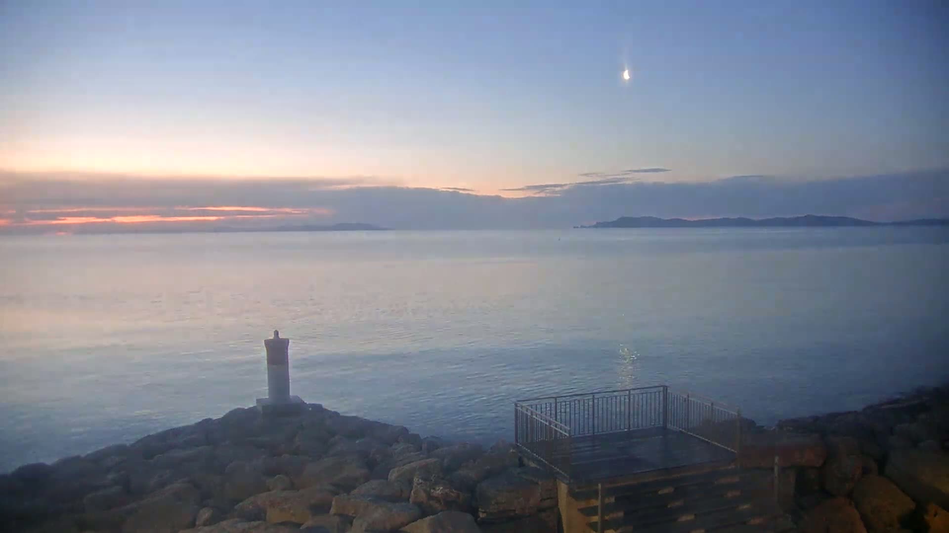 A calm sea under a twilight sky with a crescent moon visible, viewed from a rocky shoreline featuring a small lighthouse and a viewing platform.