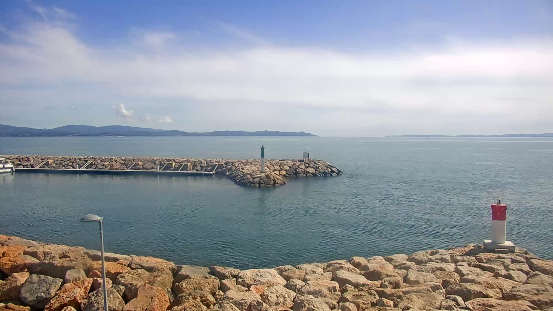 A calm, sunny day shows a rocky harbor with a small boat docked, a breakwater extending into a body of water, and distant mountains under a partly cloudy sky.