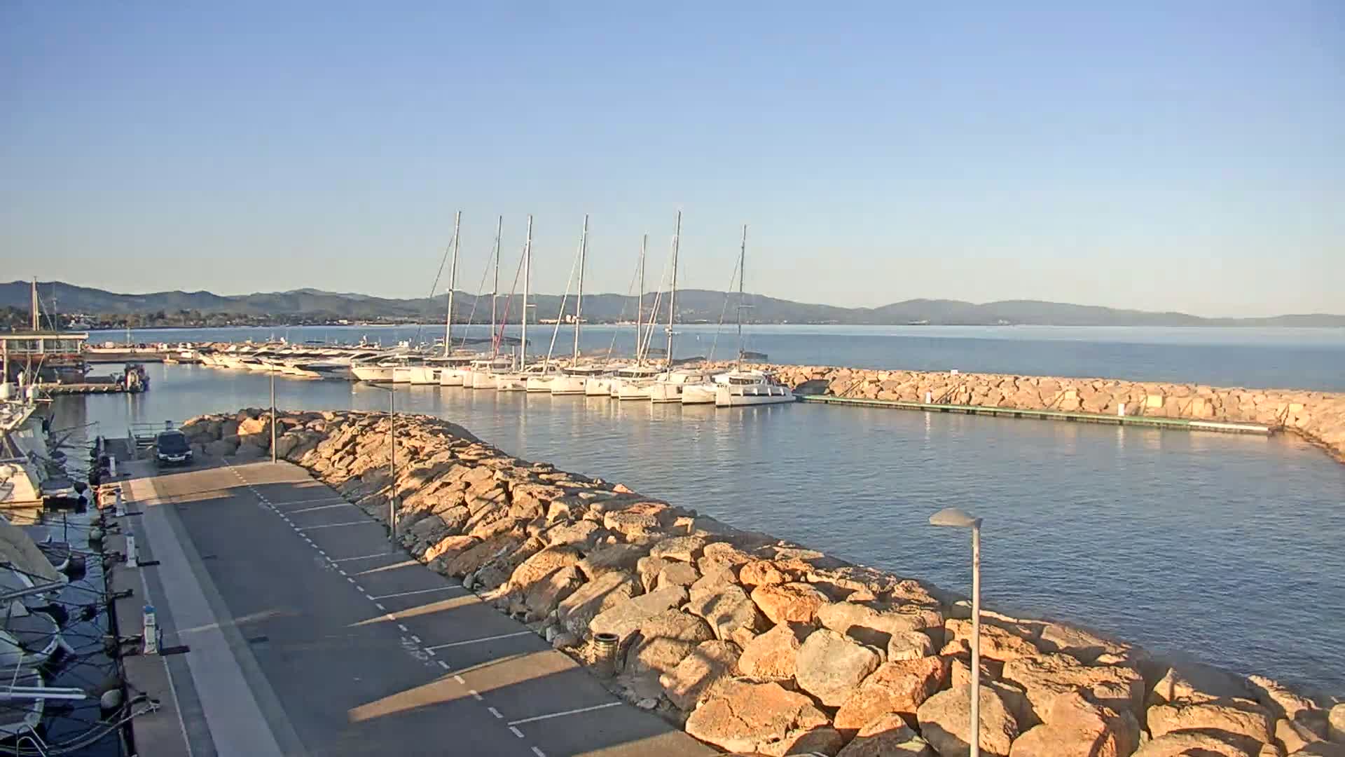 A marina filled with sailboats and catamarans is situated on calm water under a clear blue sky, protected by a rock wall and a paved area with parking.