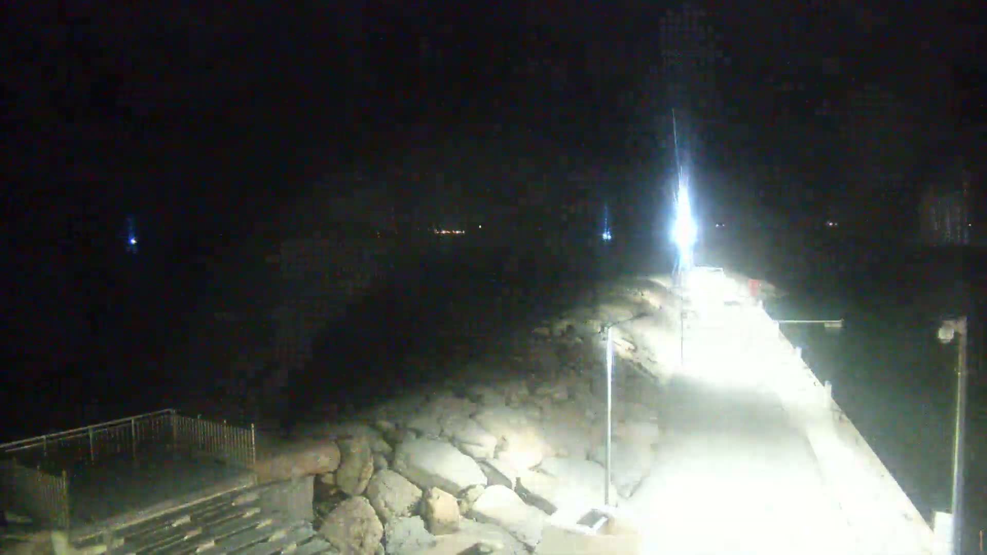 A nighttime view shows a brightly lit paved walkway along a rocky shoreline, with dark water visible beyond.