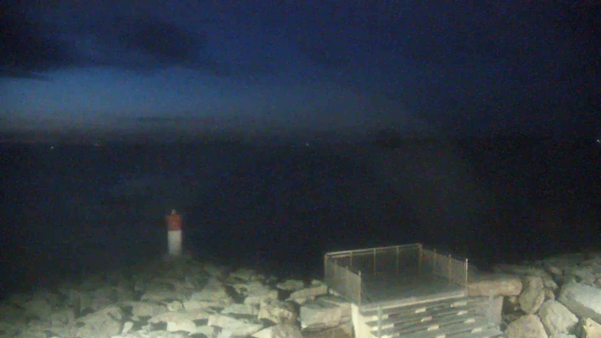 A nighttime view of a dark body of water, a rocky shoreline with a small metal structure, and a navigational marker.