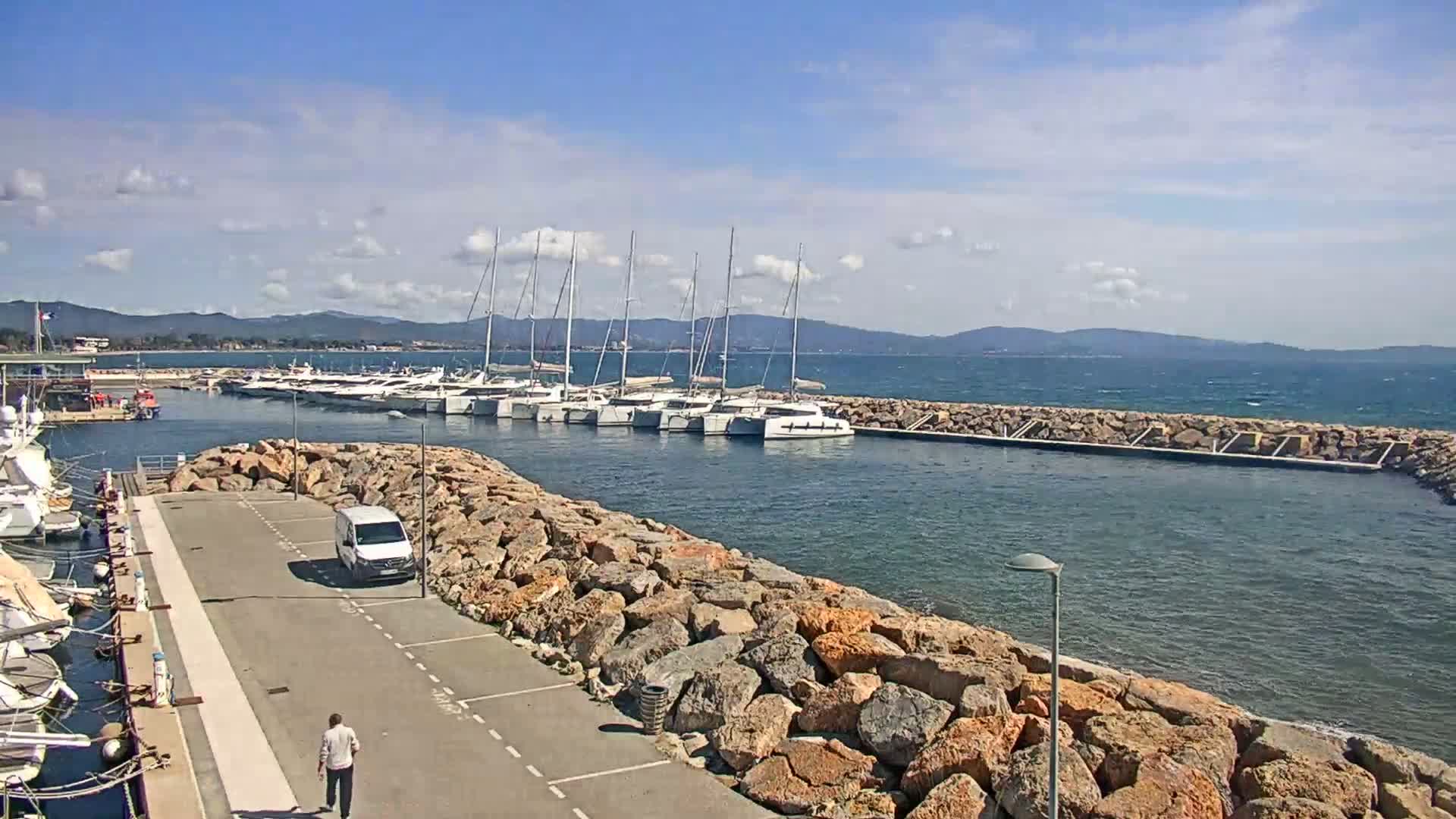 A marina filled with sailboats and yachts is partially sheltered by a rocky breakwater on a sunny day with some clouds.