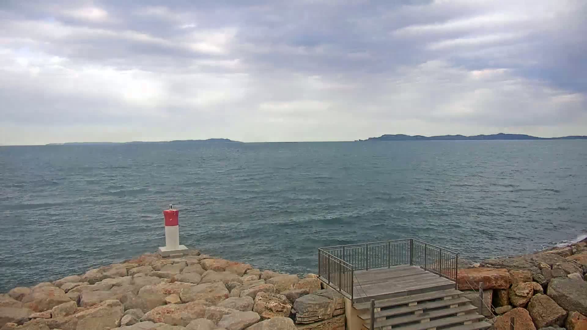 A rocky shoreline with a small viewing platform overlooks a calm sea under a cloudy sky, a distant landmass visible on the horizon.