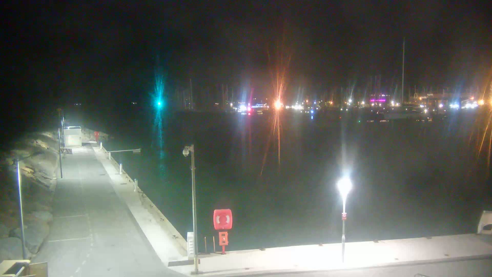 A nighttime view of a harbor with boats docked, illuminated by lights along a paved walkway.