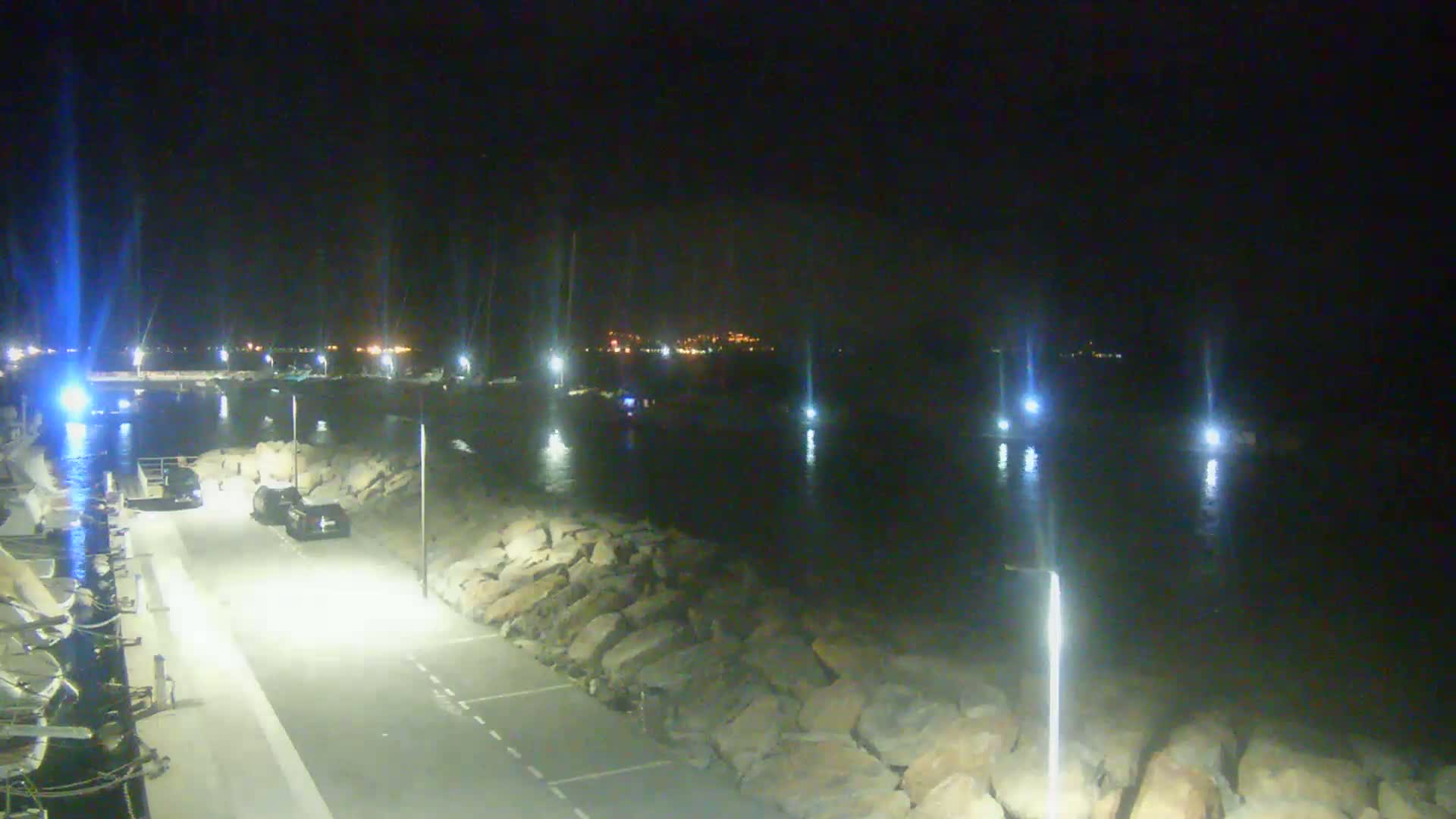 A nighttime view of a harbor with several boats moored, a rocky shoreline, and a few cars parked in a lot illuminated by bright lights.