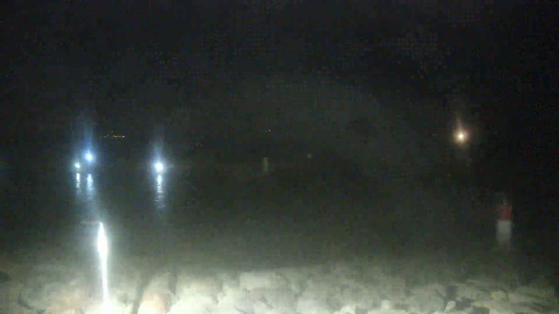 A nighttime, low-light view of a rocky shoreline with two sets of vehicle headlights visible on the water in the distance.