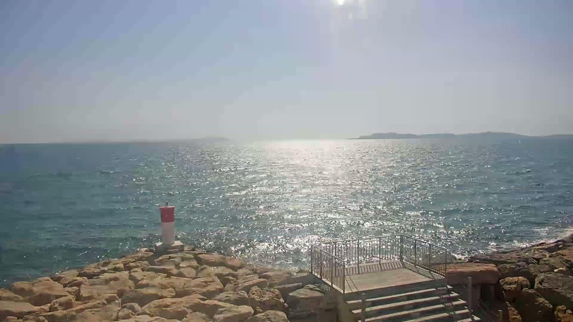 A rocky shoreline with a small wooden deck and stairs overlooks a calm, sunlit sea on a clear day.
