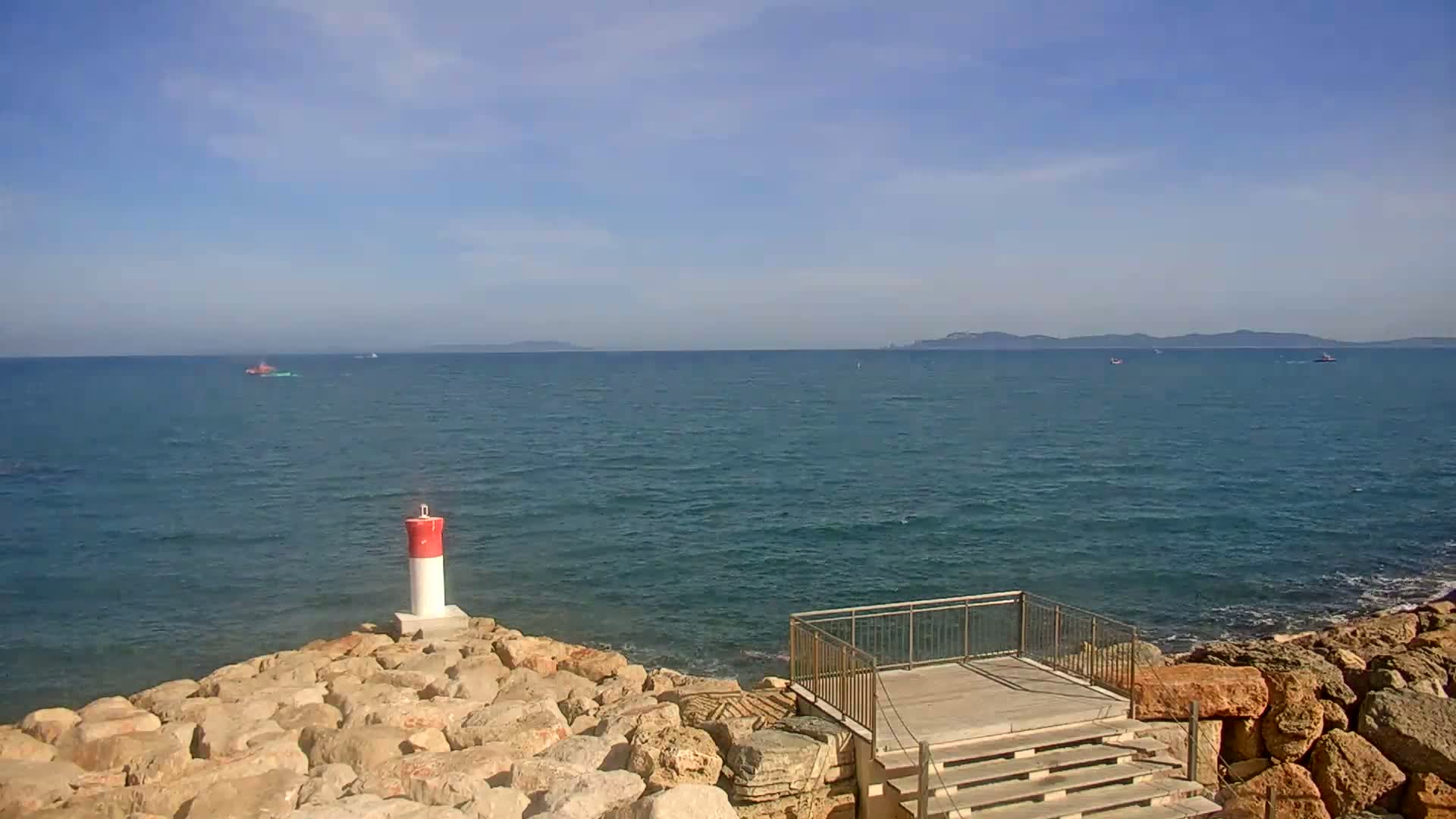 A rocky shoreline with a small wooden platform and railing overlooks a calm, blue sea under a mostly clear sky, with a few distant boats and islands visible on the horizon.