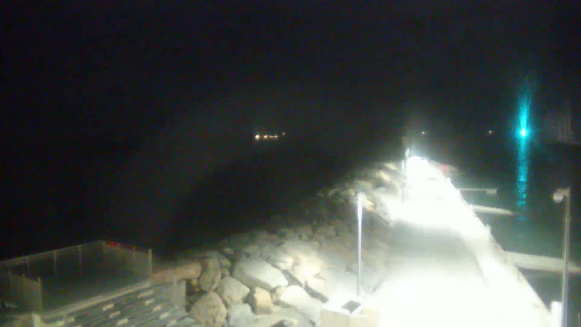 A brightly lit walkway extends from a rocky shoreline at night over dark water, with lights faintly visible in the distance.