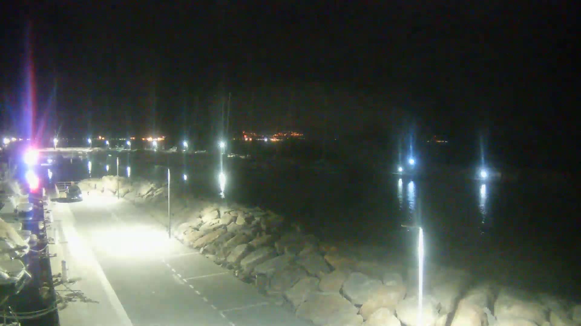 A nighttime view of a harbor, illuminated by lights along a paved walkway beside a rocky seawall, with boats visible in the dark water.