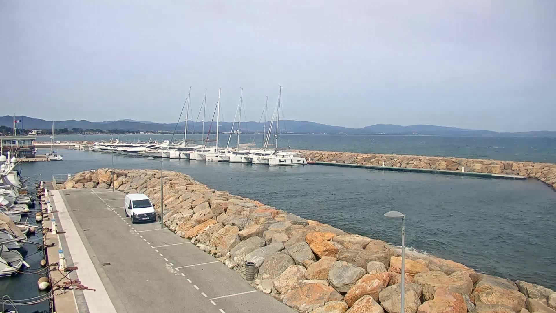 A marina with numerous sailboats and a white van parked beside a rocky breakwater on a partly cloudy day with a slightly choppy sea.