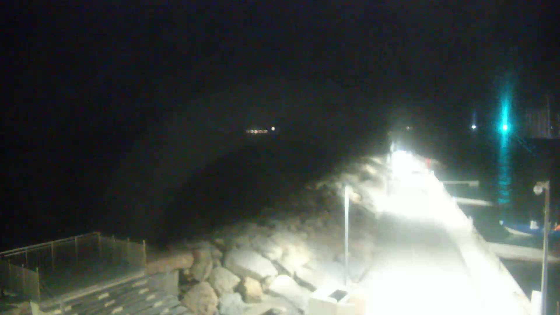 A brightly lit walkway next to a rocky shoreline at night, with boats moored in the dark water.