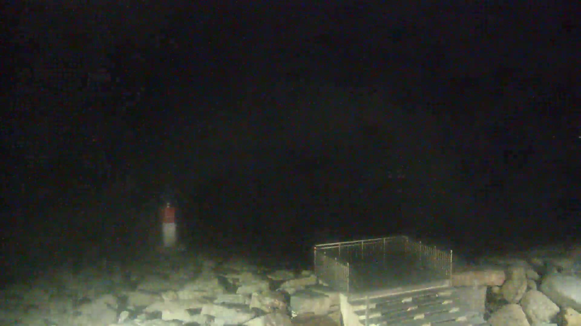 A dark, nighttime scene shows a rocky shoreline with a metal railing and a distant, indistinct marker.