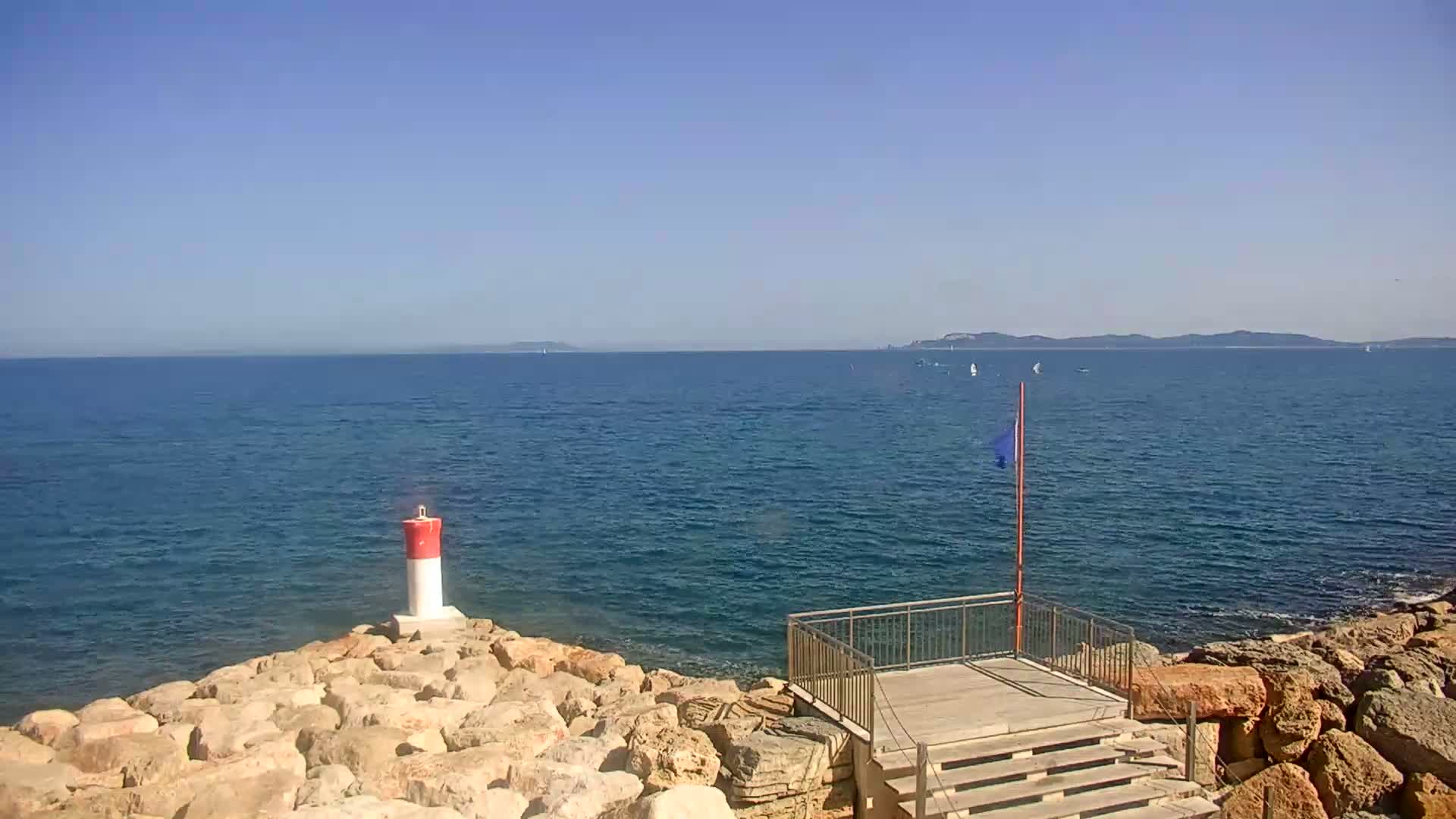 A rocky shoreline with a small wooden platform and steps overlooks a calm, blue sea on a sunny day, with a red and white marker buoy and distant land visible.