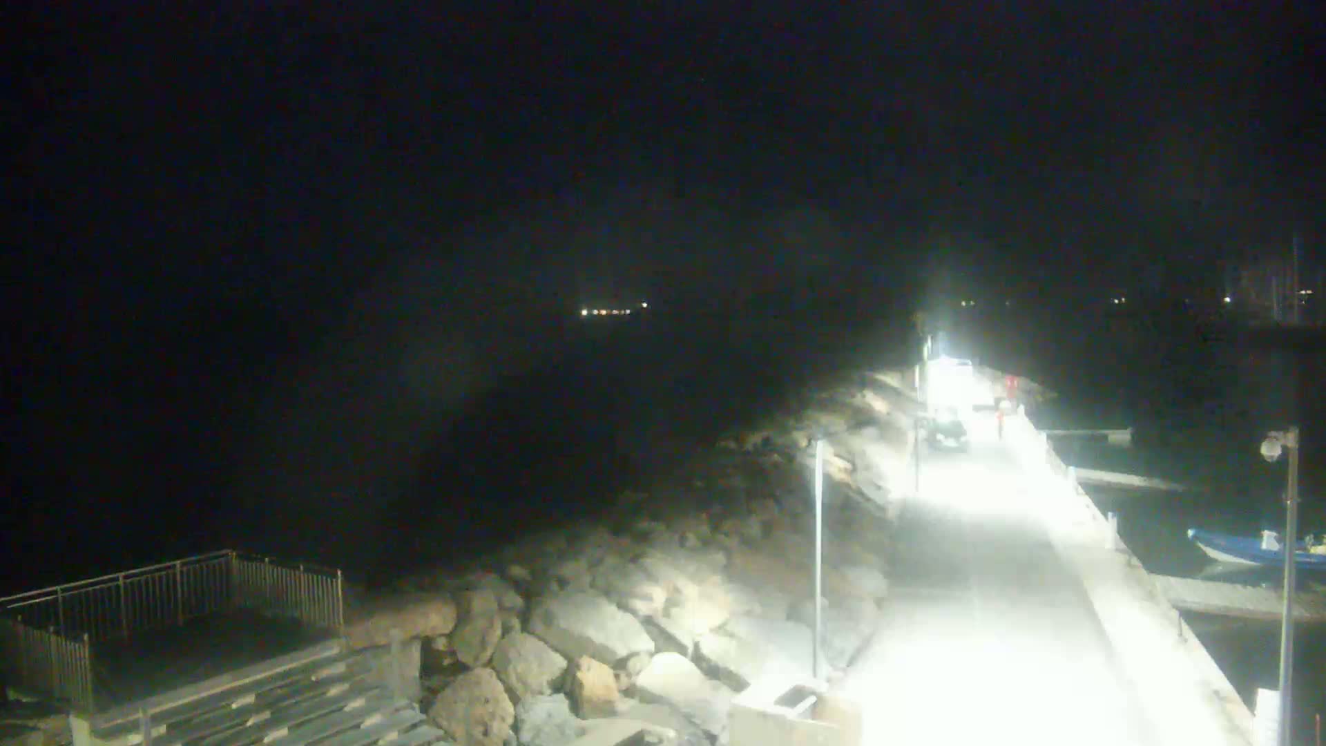 A nighttime, brightly lit paved walkway along a rocky shoreline extends toward a dark body of water, with a few small boats visible in the distance.