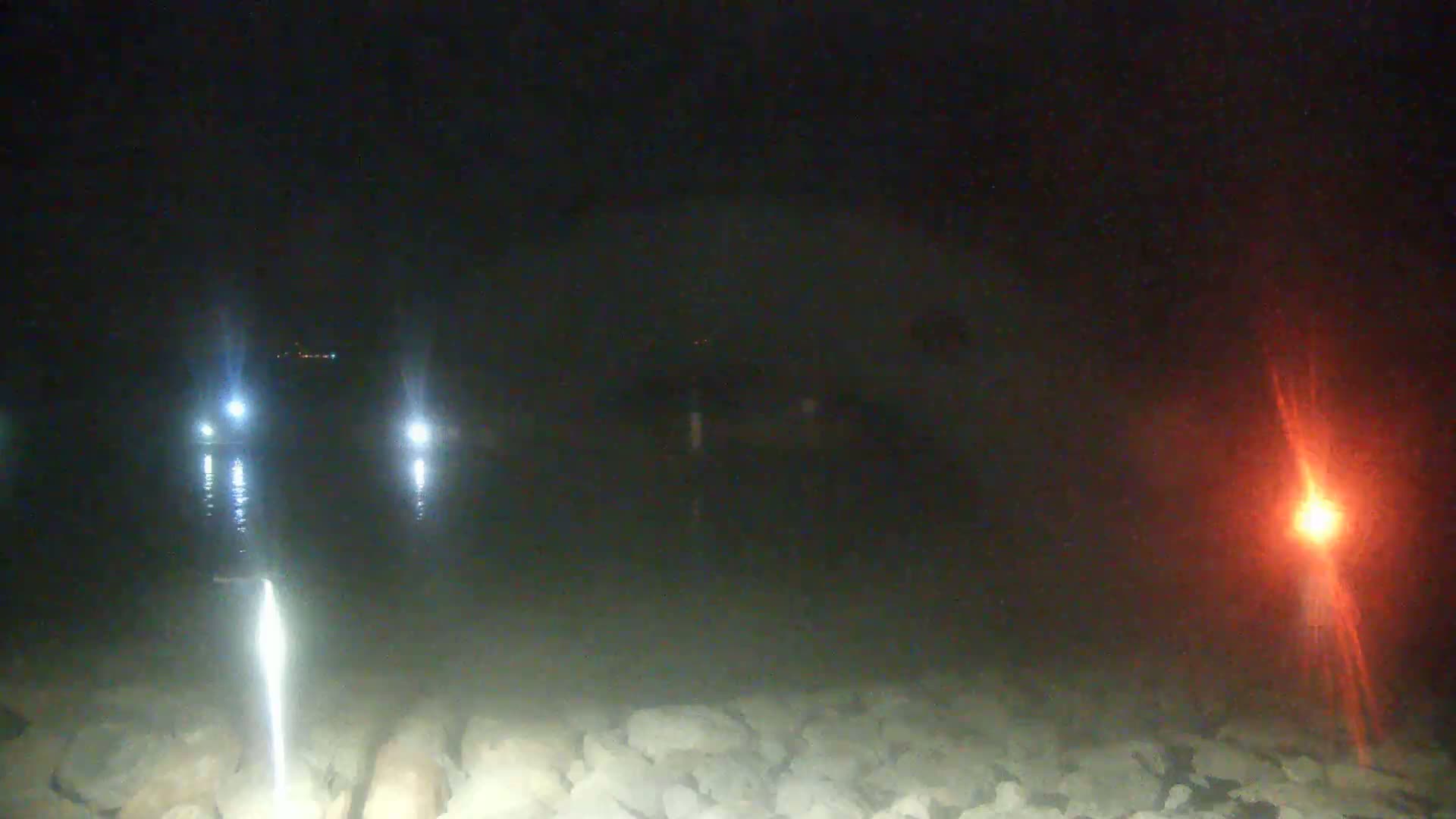 At night, bright lights illuminate a dark body of water reflecting off a rocky shore with a distant red light visible.