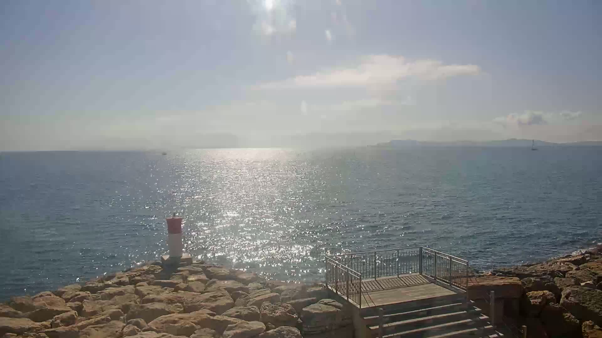 A rocky shoreline with a small wooden platform and steps overlooks a calm, sunlit sea under a mostly clear sky.