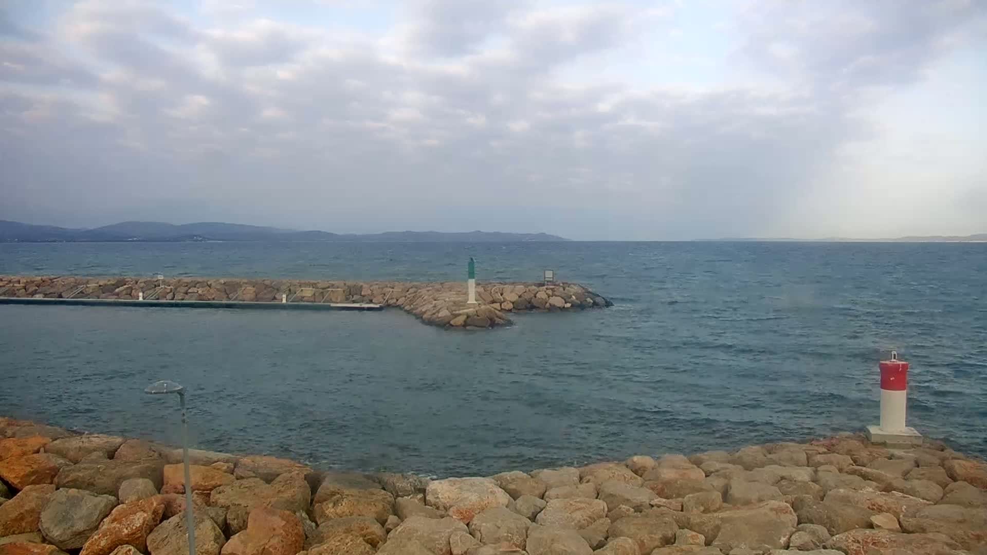 A rocky breakwater extends into a wavy sea under a cloudy sky, with distant mountains visible on the horizon.