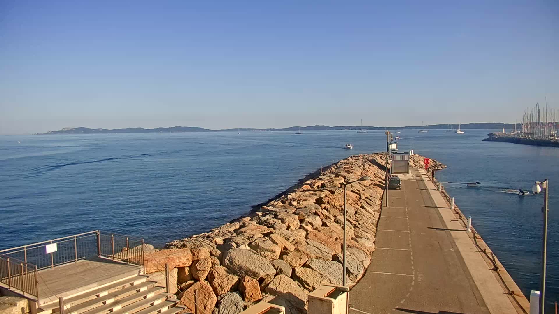 A rocky breakwater extends into a calm blue sea under a clear blue sky, with a distant coastline visible and a few boats on the water.