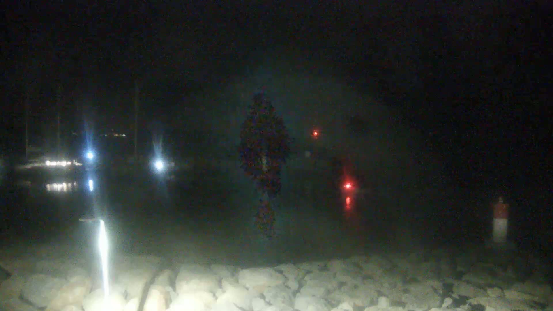 A dark, nighttime scene shows a blurry figure standing near a body of water with several lights visible in the distance and a rocky shoreline in the foreground.
