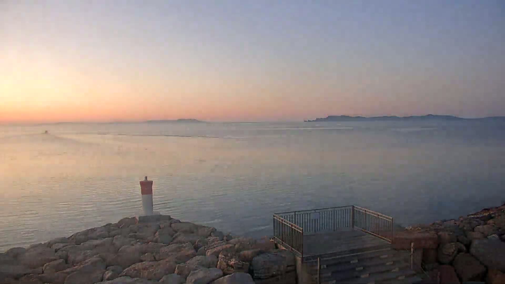 A calm, pink-hued sunrise over a body of water, viewed from a rocky shore with a small lighthouse and a viewing platform.