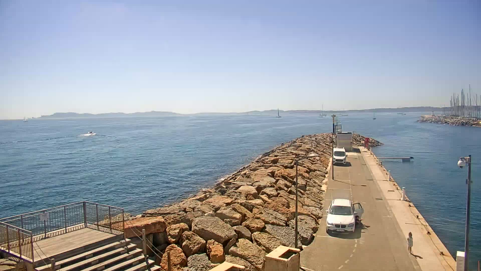 A rocky breakwater extends into a calm, blue sea under a clear, sunny sky, with a road and several cars visible on the breakwater and distant boats and islands in view.