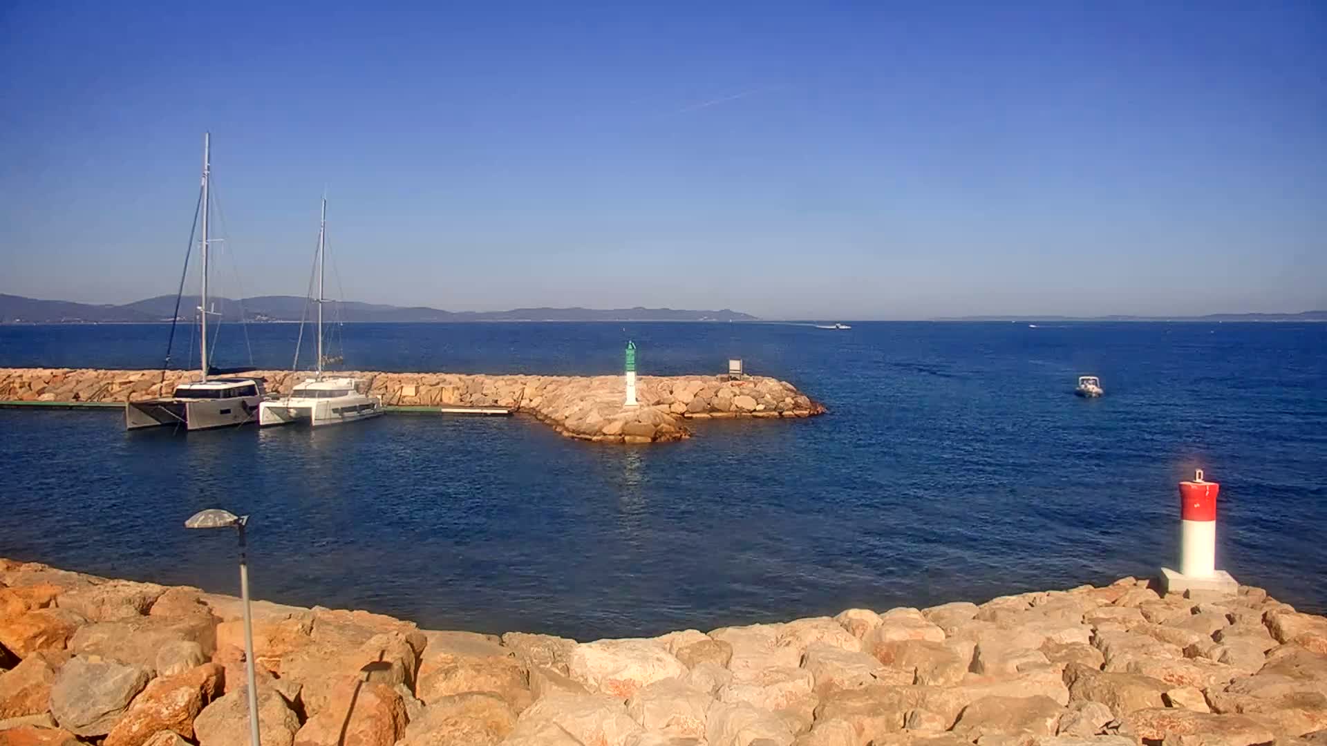 Two sailboats are moored in a calm, clear blue harbor, partially enclosed by a rocky breakwater, with a small lighthouse visible in the distance.